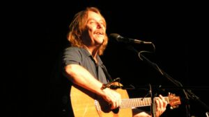 Male musician playing an acoustic guitar on stage, singing into a microphone against a dark background.