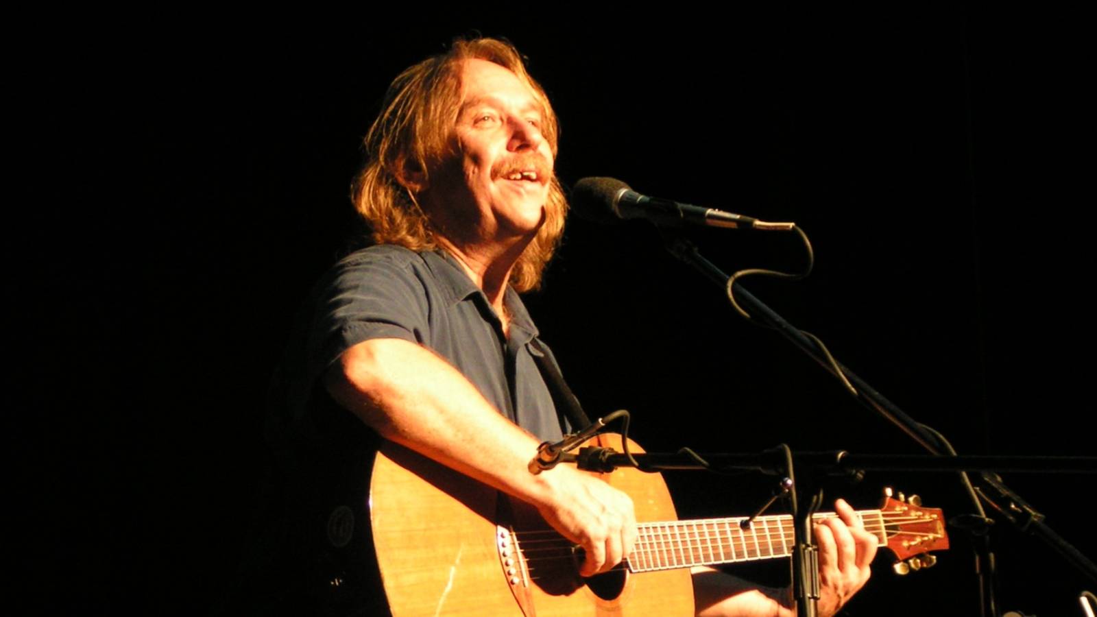 Male musician playing an acoustic guitar on stage, singing into a microphone against a dark background.