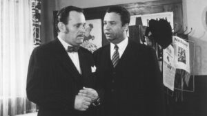 Two men in dark suits speak privately in a room, one wearing a bow tie and pocket square in a vintage setting with posters in the background.