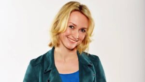 Smiling blonde woman wearing a teal jacket and blue top, posed against a light background.
