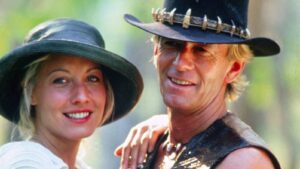 Smiling couple posing outdoors, each wearing a wide-brim hat; woman in white and man in a sleeveless shirt.