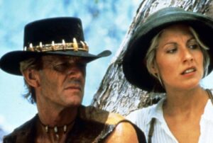 Two adults in a Western setting: a man in a black hat with a bullet necklace and a woman in a wide-brimmed hat looking to the side.