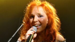 Female singer with orange hair performing on stage, holding a microphone and smiling into the mic.