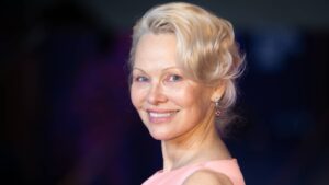 Portrait of a smiling blonde woman in a pink dress with pearl earrings against a dark blurred backdrop.