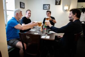 Four men celebrate with a beer toast around a table in a casual restaurant filled with natural light.