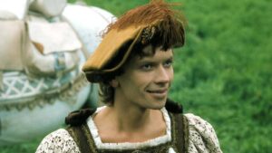 Young man wearing a brown Renaissance-style hat and lace shirt, standing in a green field with a horse in the background.