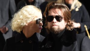 Blonde woman and brown-haired man in black coats wearing dark sunglasses outdoors, facing each other.