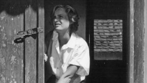 Black-and-white photo of a young woman in a white shirt peering through a weathered wooden door with a metal latch, looking anxious or fearful.