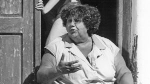 Older woman with short, curly hair sits at a doorway, gesturing with her hand as if speaking.