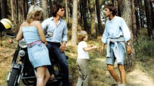 Four people stand in a pine forest beside a motorcycle, with a young boy shaking hands with a man as others look on.