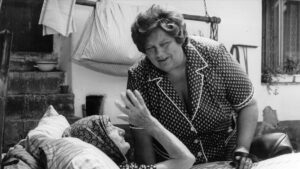 Black-and-white photo of an older woman in a patterned dress leaning toward a person lying under blankets on a porch, with clothes hanging nearby.