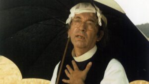 Older man wearing a clerical collar and white headwrap stands outdoors under a large black umbrella, hand resting on his chest.