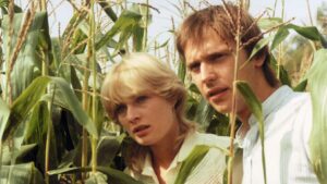 A young blond woman and a man hide among tall corn stalks, looking off-camera with wary expressions. (informative)