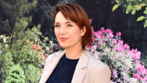 Portrait of a woman with short auburn hair, wearing a beige blazer, in a garden with pink flowers behind her.