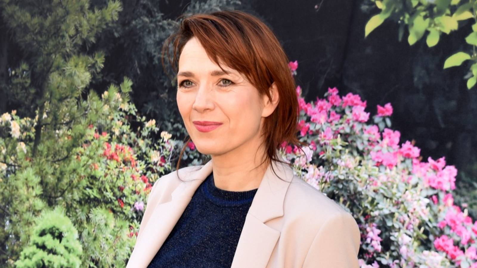 Portrait of a woman with short auburn hair, wearing a beige blazer, in a garden with pink flowers behind her.