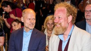 Two men in the front row at a formal event, talking to each other, with a photographer and other guests in the background.