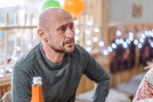 Man with a shaved head and stubble sits at a table, an orange drink in the foreground, with festive blurred lights in the background.