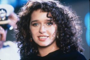 Close-up portrait of a woman with dark, curly hair and blue eyes, smiling softly at the camera, wearing a black top.