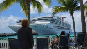Cruise ship Carnival Sunshine docked at a tropical port, seen from a row of lounge chairs under palm trees.