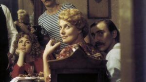 Group of people seated around a dining table at a social gathering; a woman with curly red hair in a red sweater rests her chin on her hand and looks at the camera.