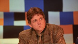 Man in a brown checked blazer and tie sits at a desk on a colorful, geometric backdrop, glancing to the side.