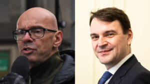 Two men side by side: left, bald man with glasses speaking into a microphone; right, man in a dark suit smiling.