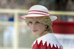 Woman wearing a white hat with red trim, pearl necklace and earrings, in a red-and-white outfit outdoors, looking over her shoulder.