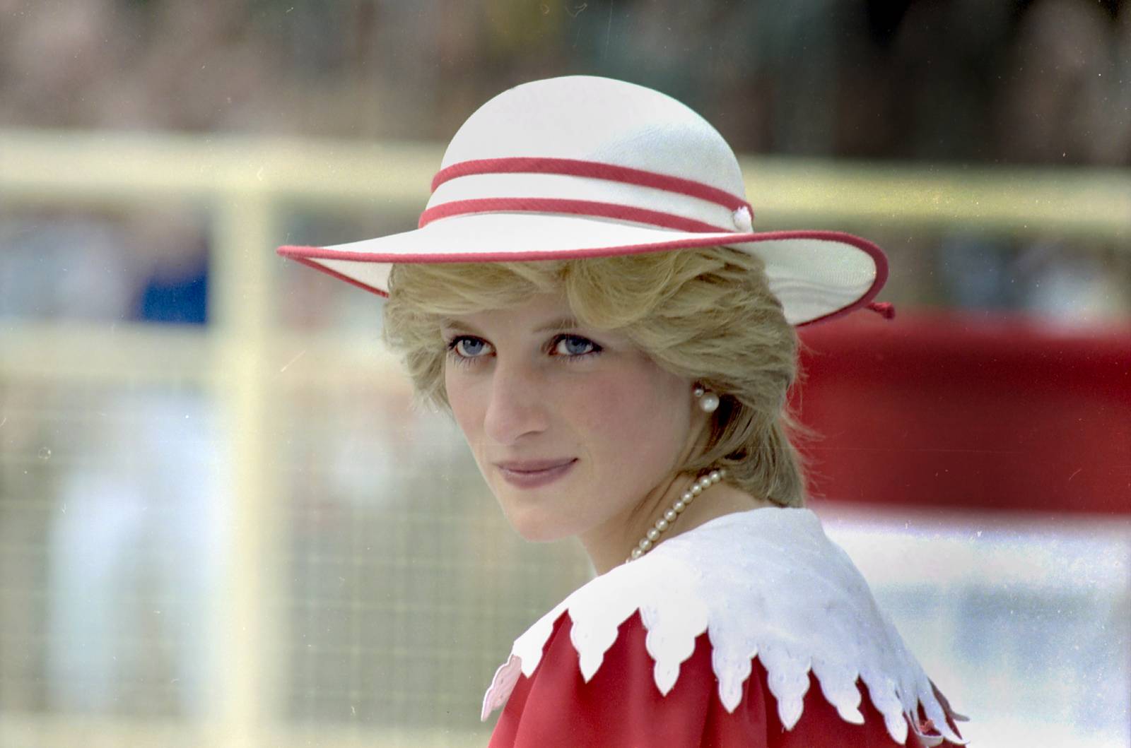 Woman wearing a white hat with red trim, pearl necklace and earrings, in a red-and-white outfit outdoors, looking over her shoulder.