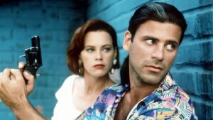 Man in a colorful tropical shirt looks to the side as a woman behind him holds a revolver, against a blue brick wall.