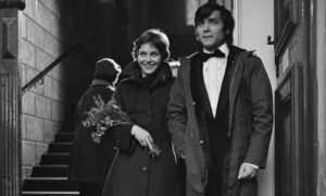 Smiling woman in a dark coat carries a bouquet as she steps out with a man in a suit and bow tie.