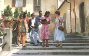 Group of casually dressed people standing on sunlit stone stairs in a narrow historic street, chatting as they face each other; a woman in a pink dress holds a notebook while another woman wears a light blue skirt and floral top with a brown purse.