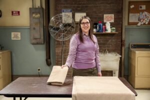 Smiling woman in a purple sweater and glasses folds beige towels on a metal table in a laundry room with brick wall behind her.